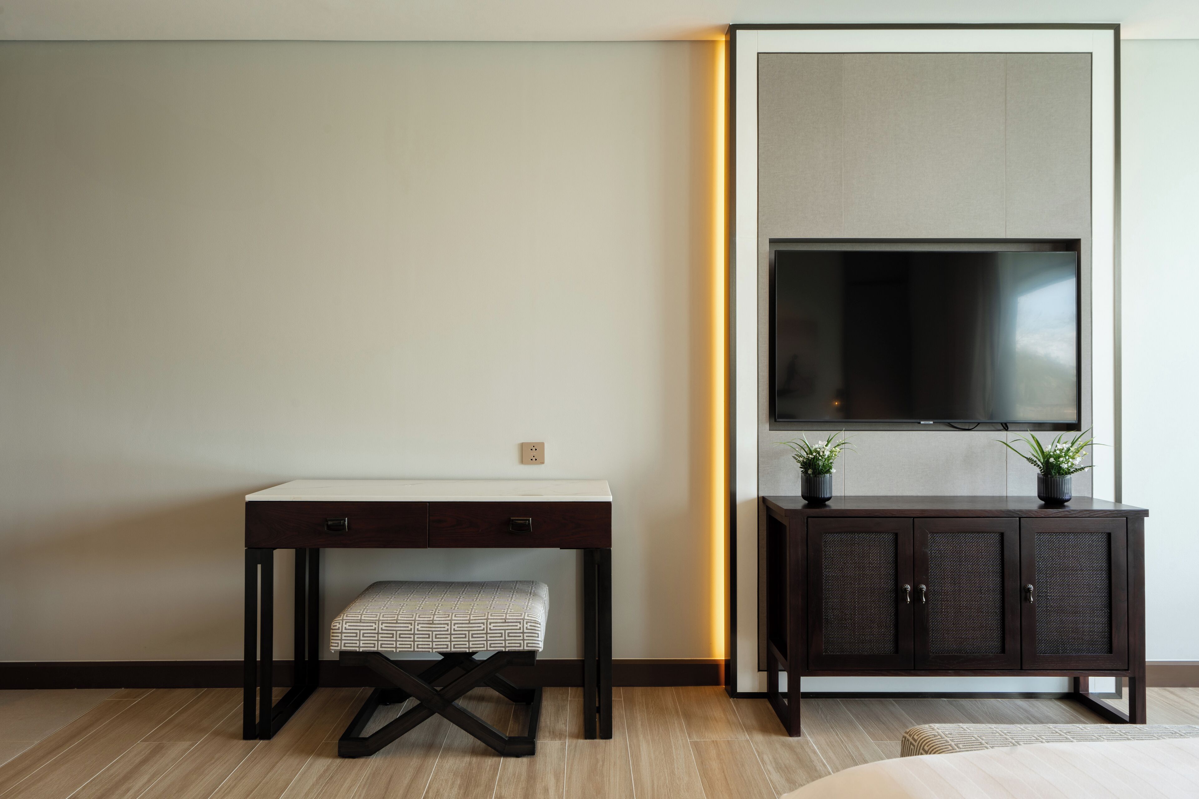 Une pièce moderne avec un bureau en bois foncé, un banc, un meuble orné de plantes et un grand téléviseur à écran plat fixé au mur.