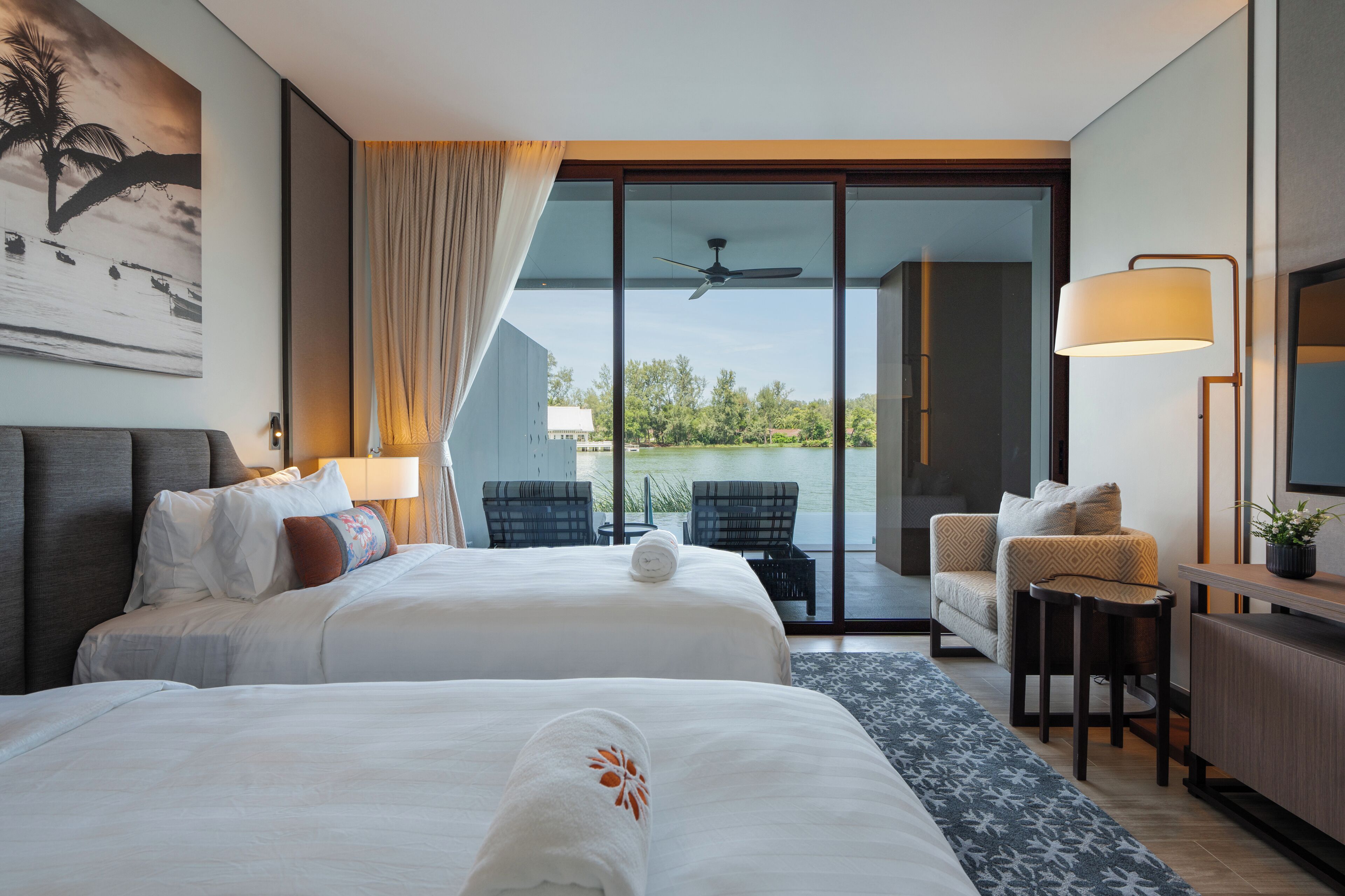 Habitación de hotel con dos camas, un escritorio, una silla, un sofá, un balcón con vistas al agua y un ventilador de techo.