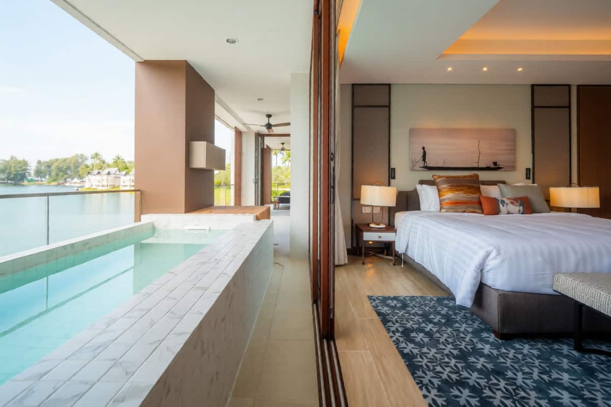 A modern hotel room with a large bed, bedside lamps, a painting, a view of a pool, and a balcony.