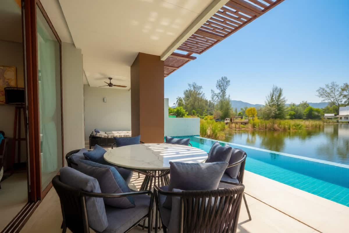 A poolside area with a round table and chairs, a ceiling fan, and a view of a lake and trees.