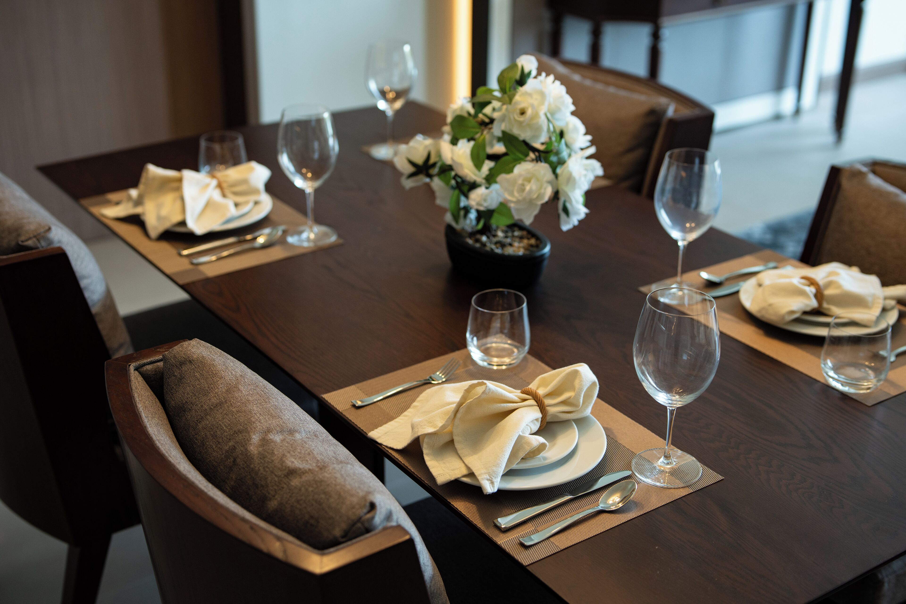 A dining table set with white napkins, silverware, and wine glasses, accompanied by a vase of white flowers.
