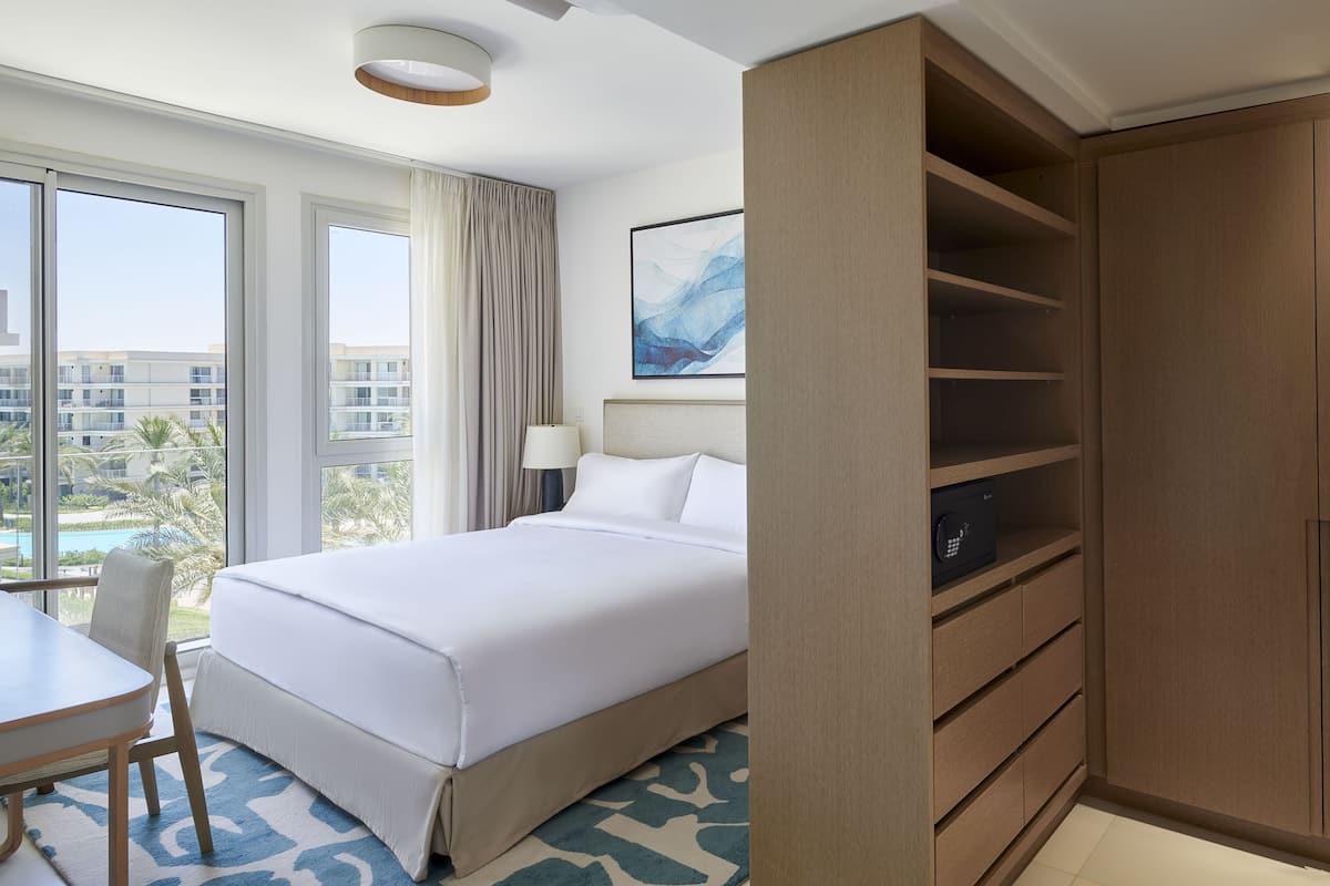 Habitación de hotel con cama, escritorio, silla, armario y vistas a una piscina y edificios.