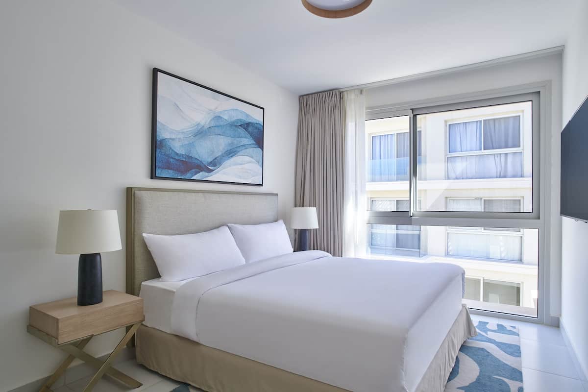 Habitación de hotel con una cama grande, dos lámparas de noche, un cuadro abstracto enmarcado, una ventana con cortinas y vistas al exterior del edificio.