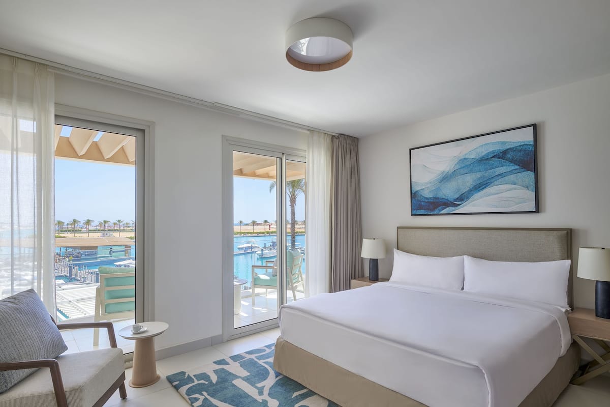 Habitación de hotel con una cama grande, vista a una piscina y palmeras, y un cuadro en la pared.