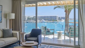 Habitación de hotel con balcón grande, sofá, silla, mesita y vista a un puerto deportivo con barcos y edificios.