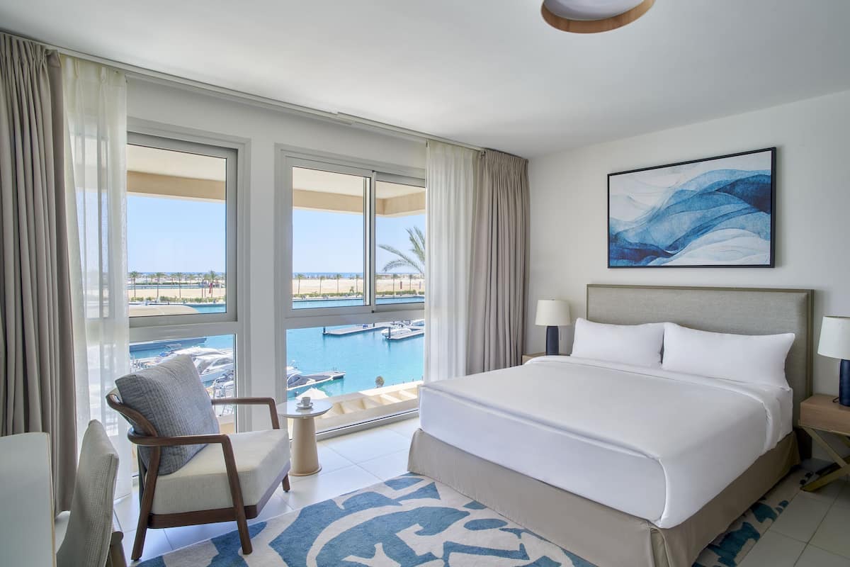 Habitación de hotel con una cama grande, una silla, una mesita y vista a un puerto deportivo.