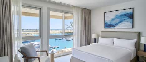 Ein Hotelzimmer mit einem großen Bett, einem Sessel, einem kleinen Tisch und Blick auf einen Yachthafen.
