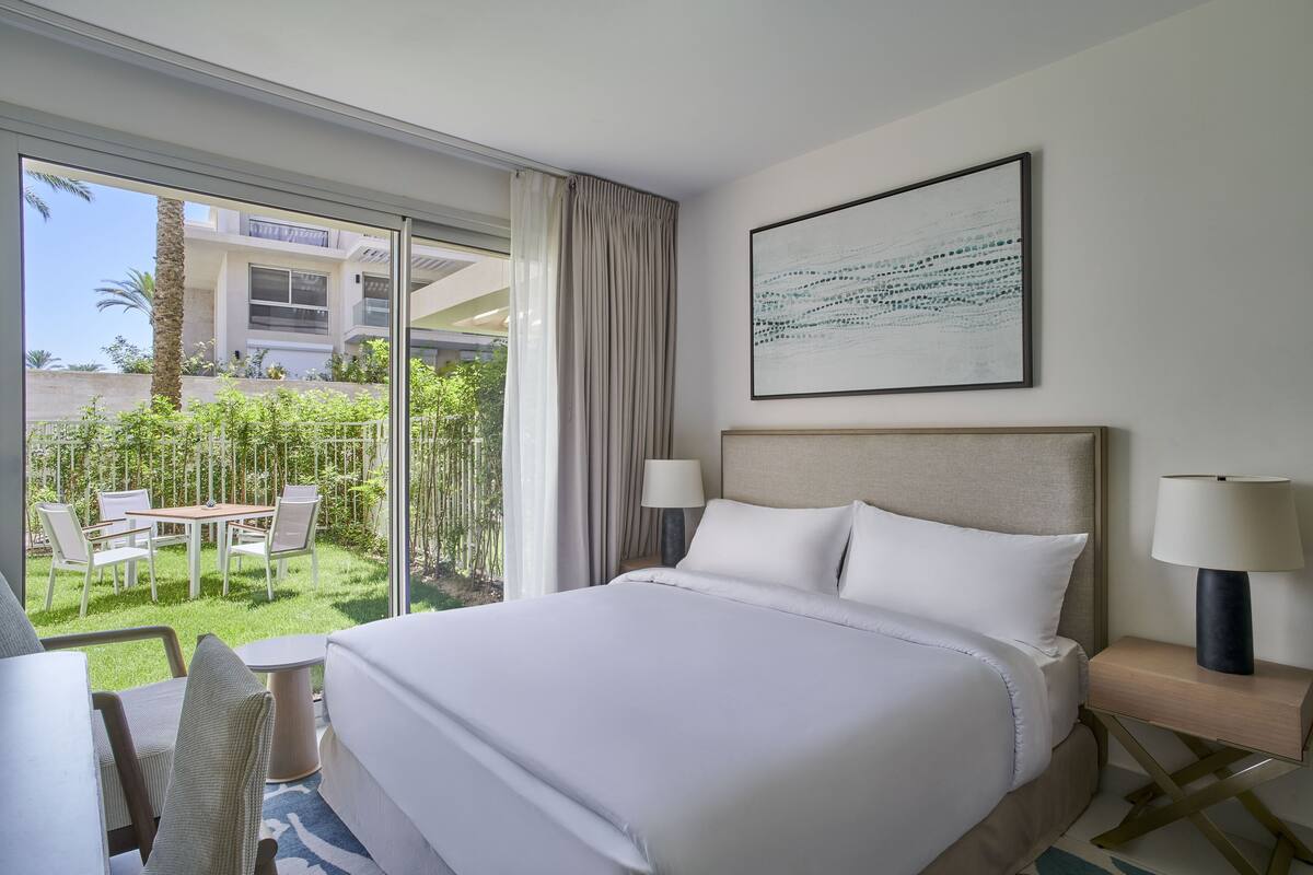 Habitación de hotel con una cama grande, un escritorio y una silla. Se ve una ventana con vistas a un edificio y un jardín.