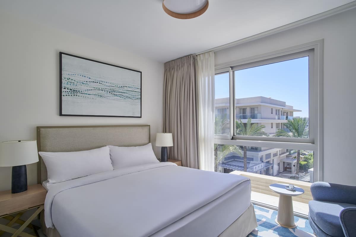 Habitación de hotel con una cama grande, dos mesitas de noche con lámparas, una pequeña mesa redonda y vistas a edificios a través de un ventanal.