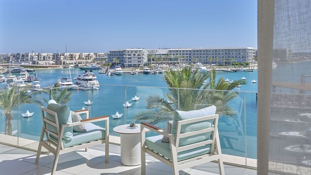 Un balcón con dos sillas, una mesita y vista a un puerto deportivo con barcos y un hotel llamado 'VIDA' al fondo.