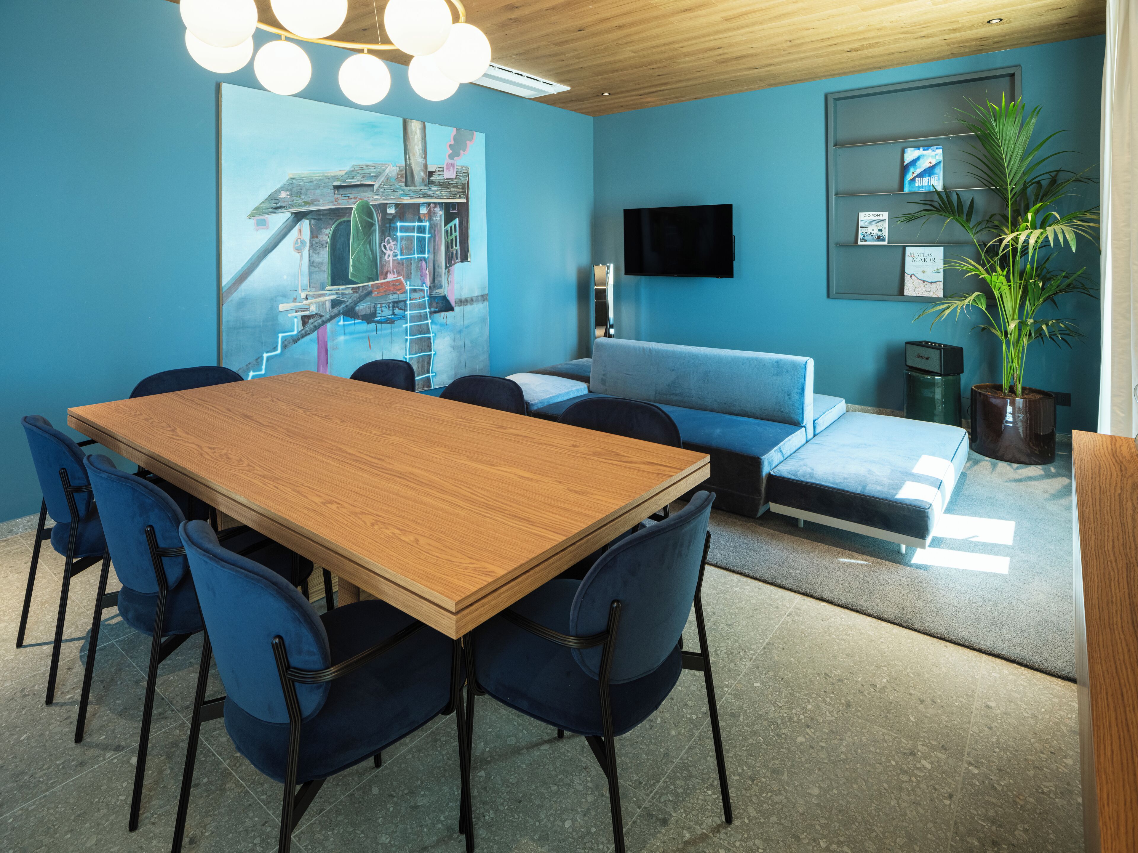 A conference room with a wooden table, blue chairs, a sofa, a TV, and a plant.