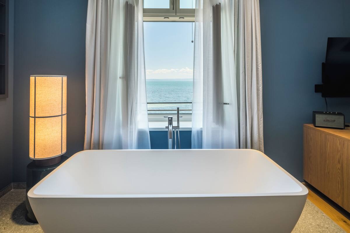 Ein modernes Badezimmer mit einer freistehenden Badewanne, einer Stehlampe und Meerblick durch ein Fenster mit transparenten Vorhängen.