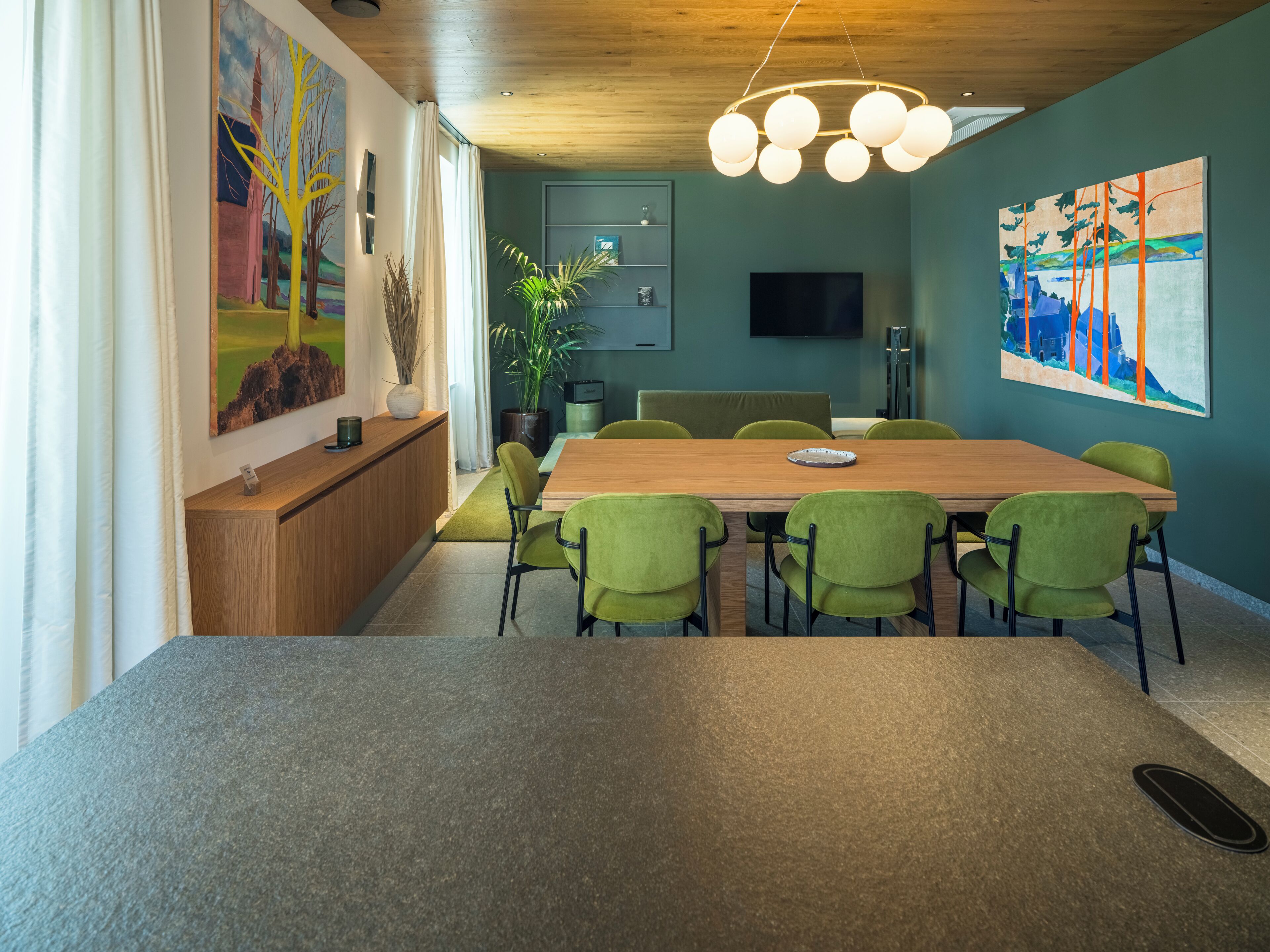 A modern meeting room with a wooden table, green chairs, a large painting, and a ceiling light fixture.