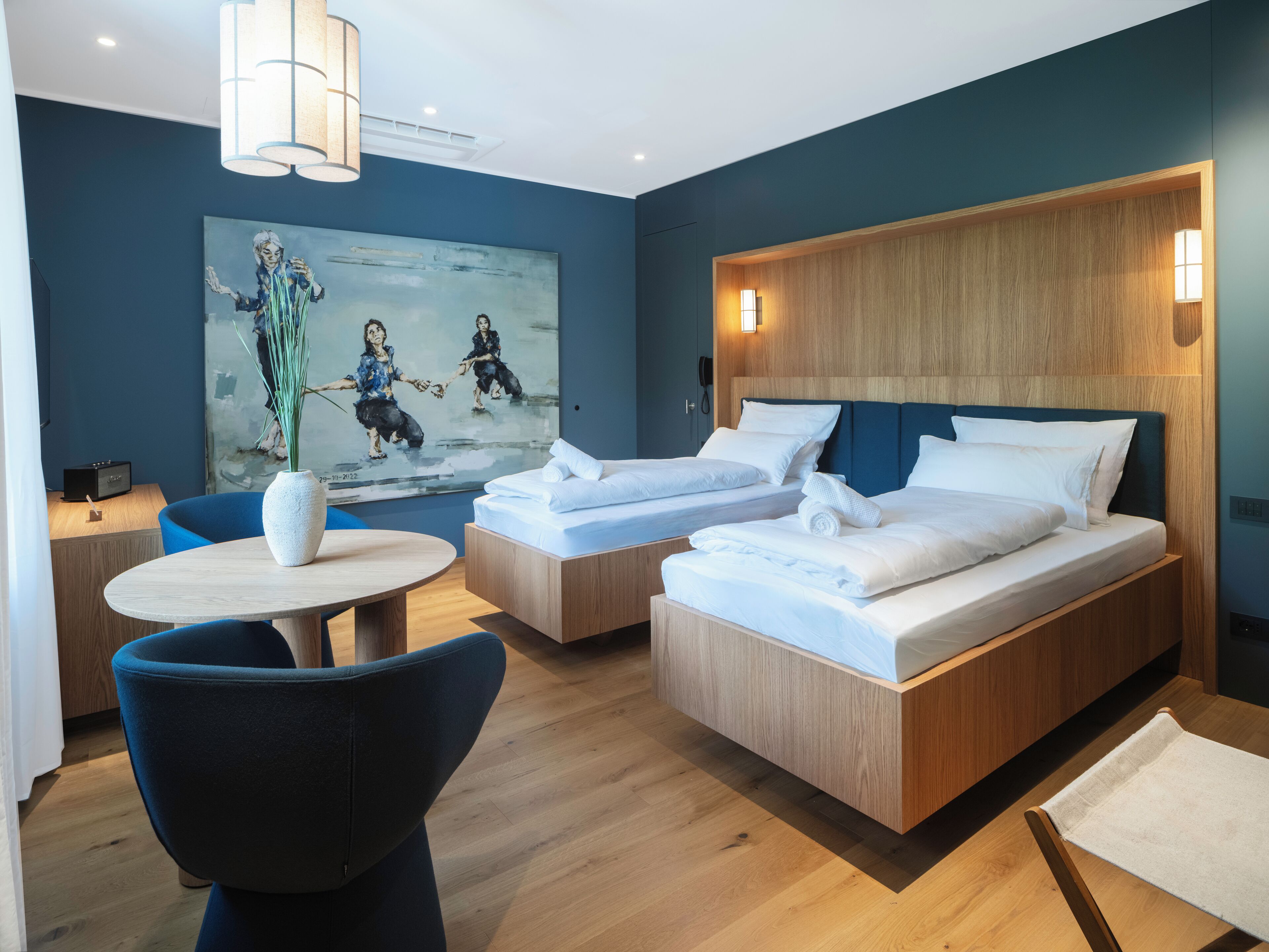 Une chambre d’hôtel moderne avec deux lits, une table ronde, une chaise, un tableau au mur et une tête de lit en bois.
