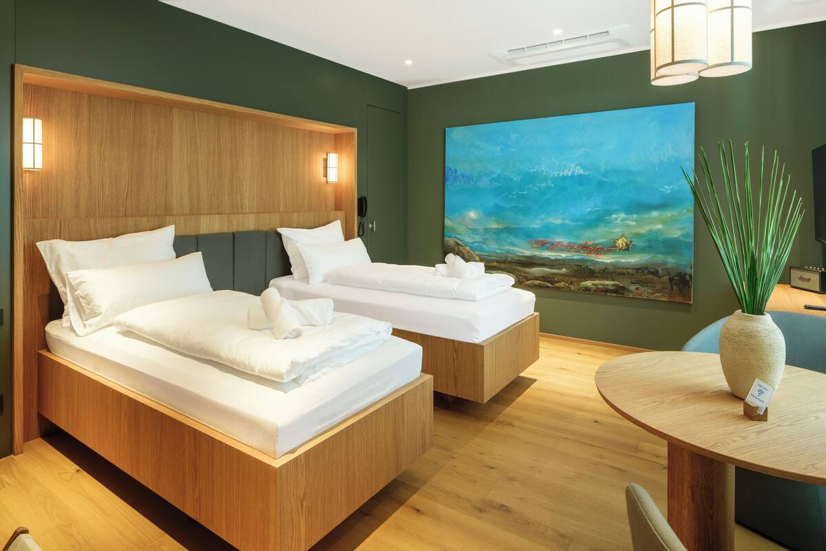 Ein modernes Hotelzimmer mit zwei Betten, einem hölzernen Kopfteil, einem Landschaftsbild und einem runden Holztisch mit einer Vase.