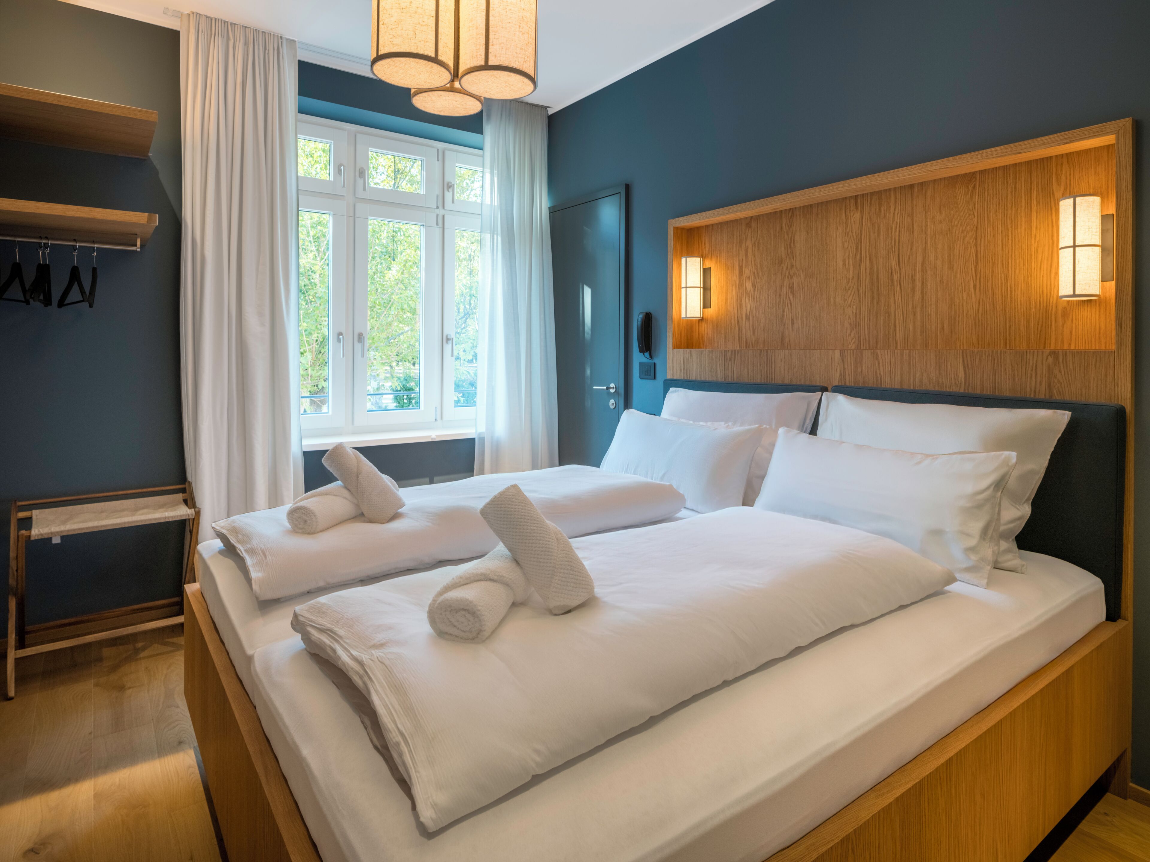 Quarto de hotel com duas camas, uma cabeceira de madeira, uma janela com cortinas, um porta-toalhas pendurado e um telefone fixado na parede.