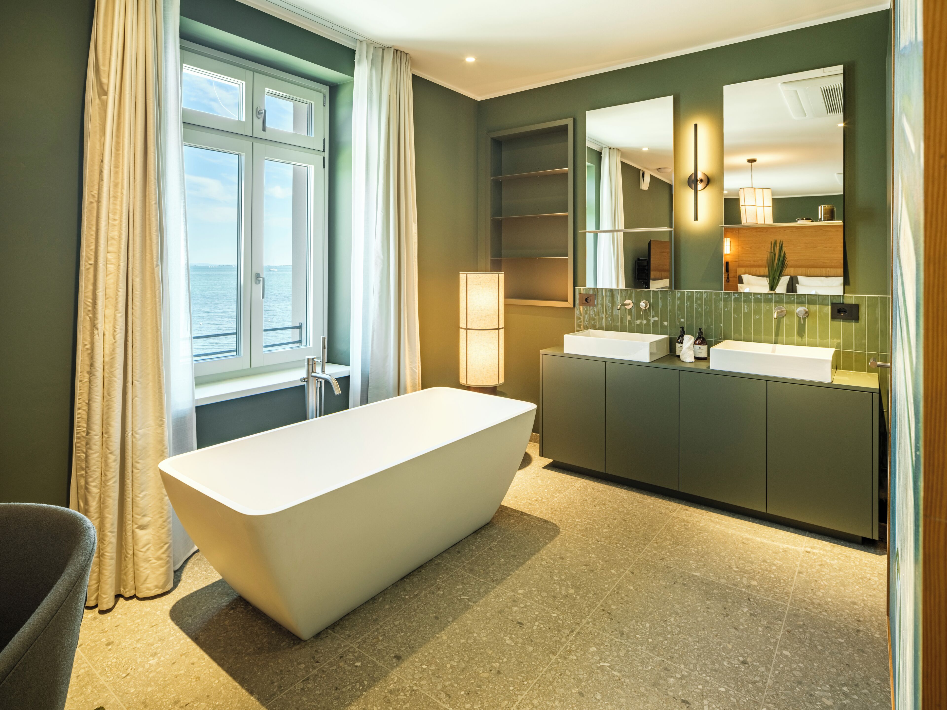 Un baño moderno con una bañera independiente, un tocador con espejo y vistas al mar a través de una ventana.