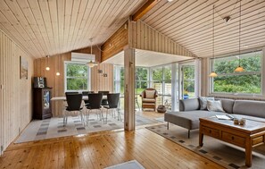 A spacious room with wooden floors and walls, a dining area with chairs and a table, a sofa, and large windows offering views of greenery. - 3 bedroom awesome home in Frederikshavn (Frederikshavn)