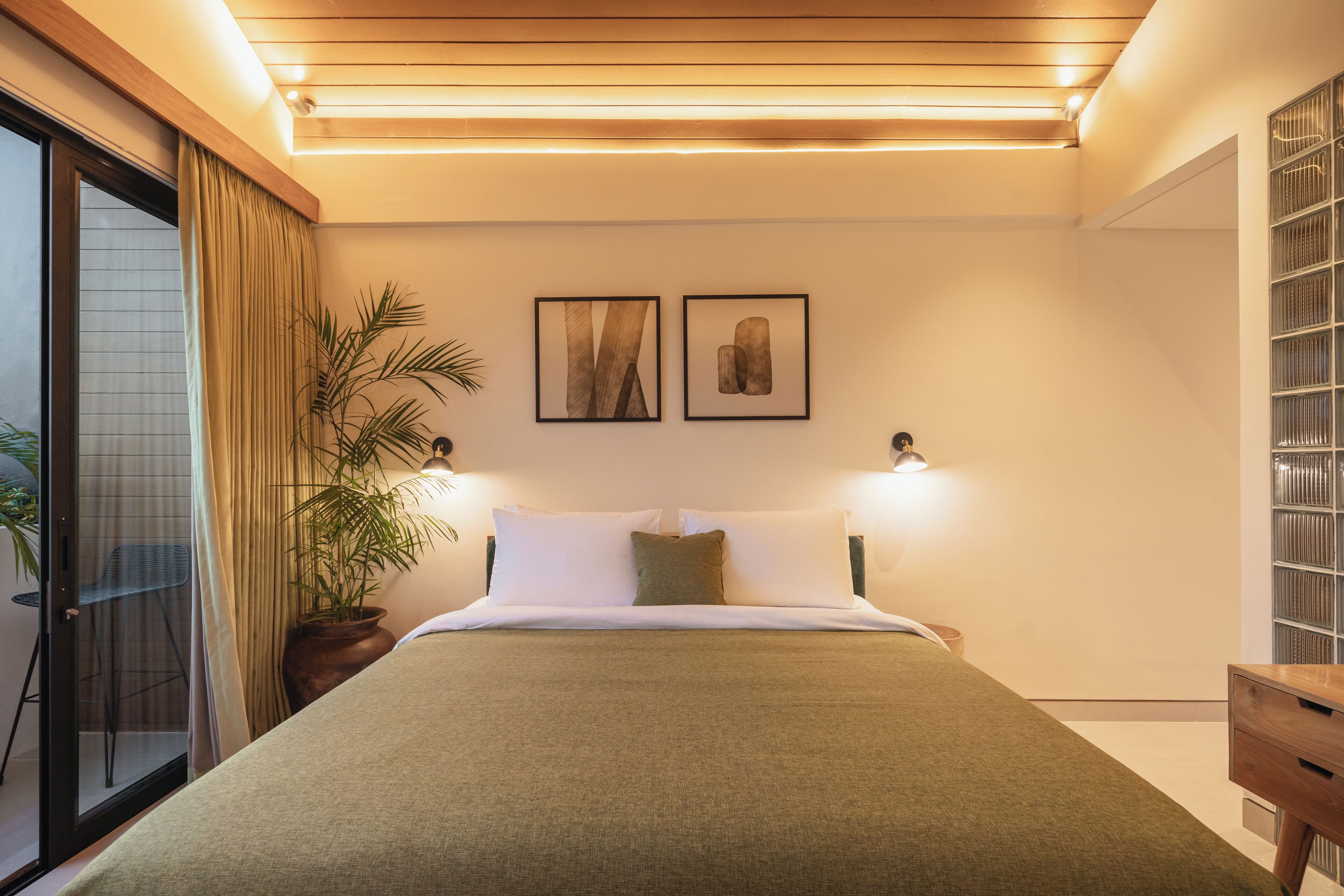Un dormitorio moderno con una cama grande, dos lámparas fijadas a la pared, una planta en maceta y un ventanal corredero que da a un balcón.
