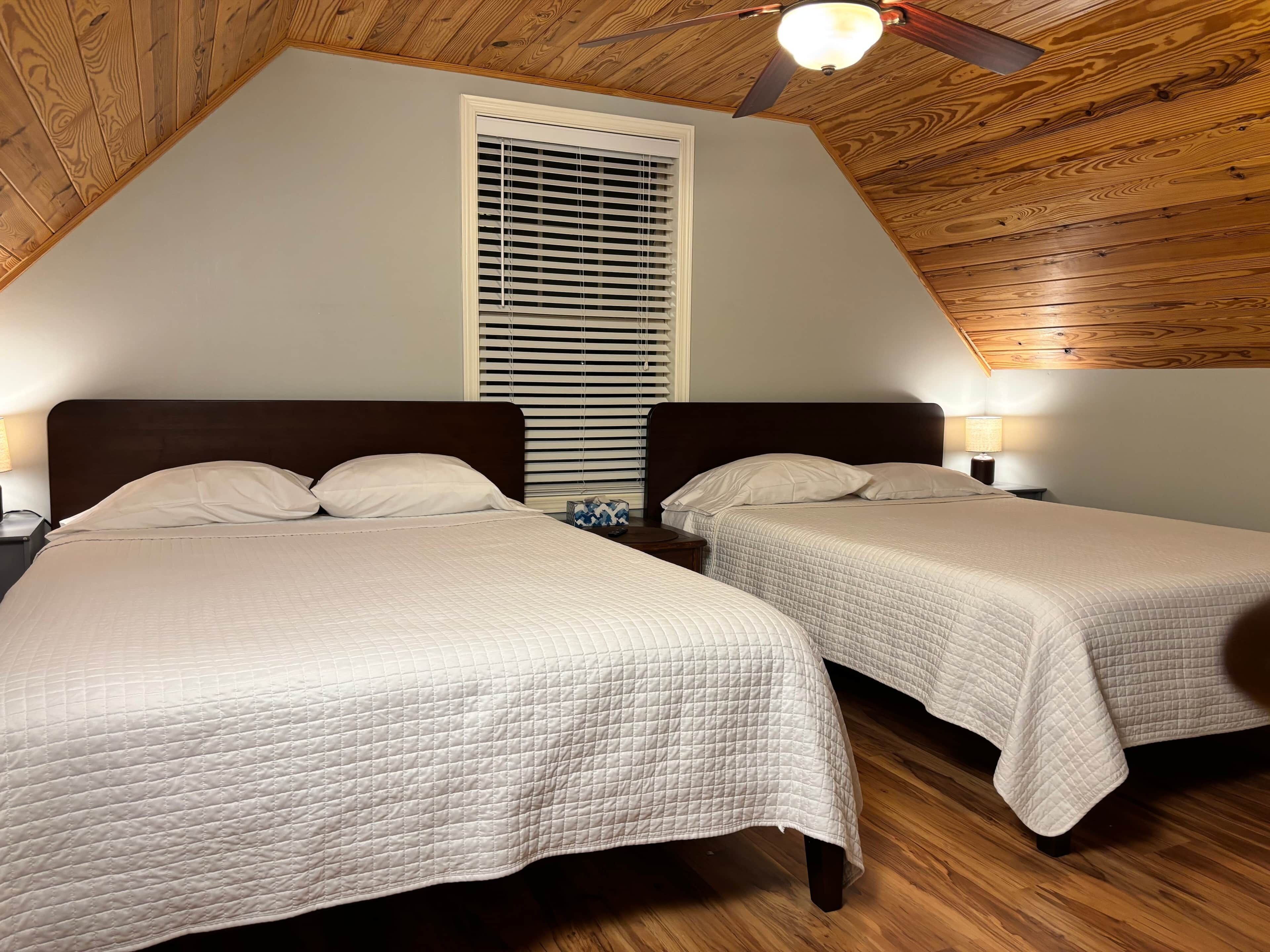 A bedroom with two beds, wooden ceiling, and a window with blinds.