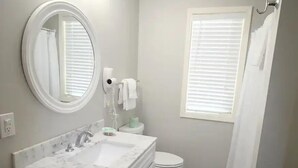 Une salle de bain avec un meuble-lavabo blanc, un miroir rond, des toilettes et une fenêtre avec des stores.
