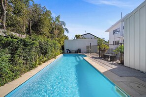 Une piscine dotée d’une terrasse en bois, entourée de verdure et bordée d’un bâtiment blanc avec un balcon.