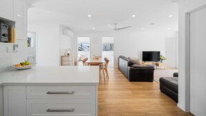 A modern kitchen with white cabinetry, a dining area with a wooden table, and a living room with a black sofa and a flat-screen TV. - The Palms in Coolum Beach - Unit A (Coolum Region)