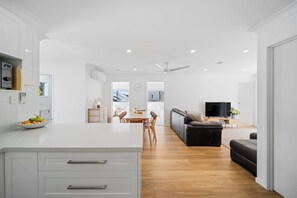 A modern kitchen with white cabinetry, a dining area with a wooden table, and a living room with a black sofa and a flat-screen TV. - The Palms in Coolum Beach - Unit A (Coolum Region)