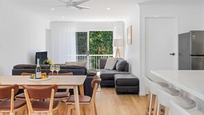 A modern living space with a dining area, a sofa, a kitchen, and a balcony with a view of greenery. - The Palms in Coolum Beach - Unit A (Coolum Region)