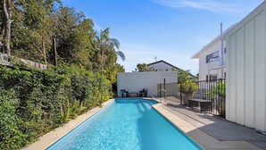 A rectangular swimming pool with a clear blue water surface, surrounded by a concrete deck. There is a white building with a balcony on the right side and a wooden fence on the left. - The Palms in Coolum Beach - Unit A (Coolum Region)