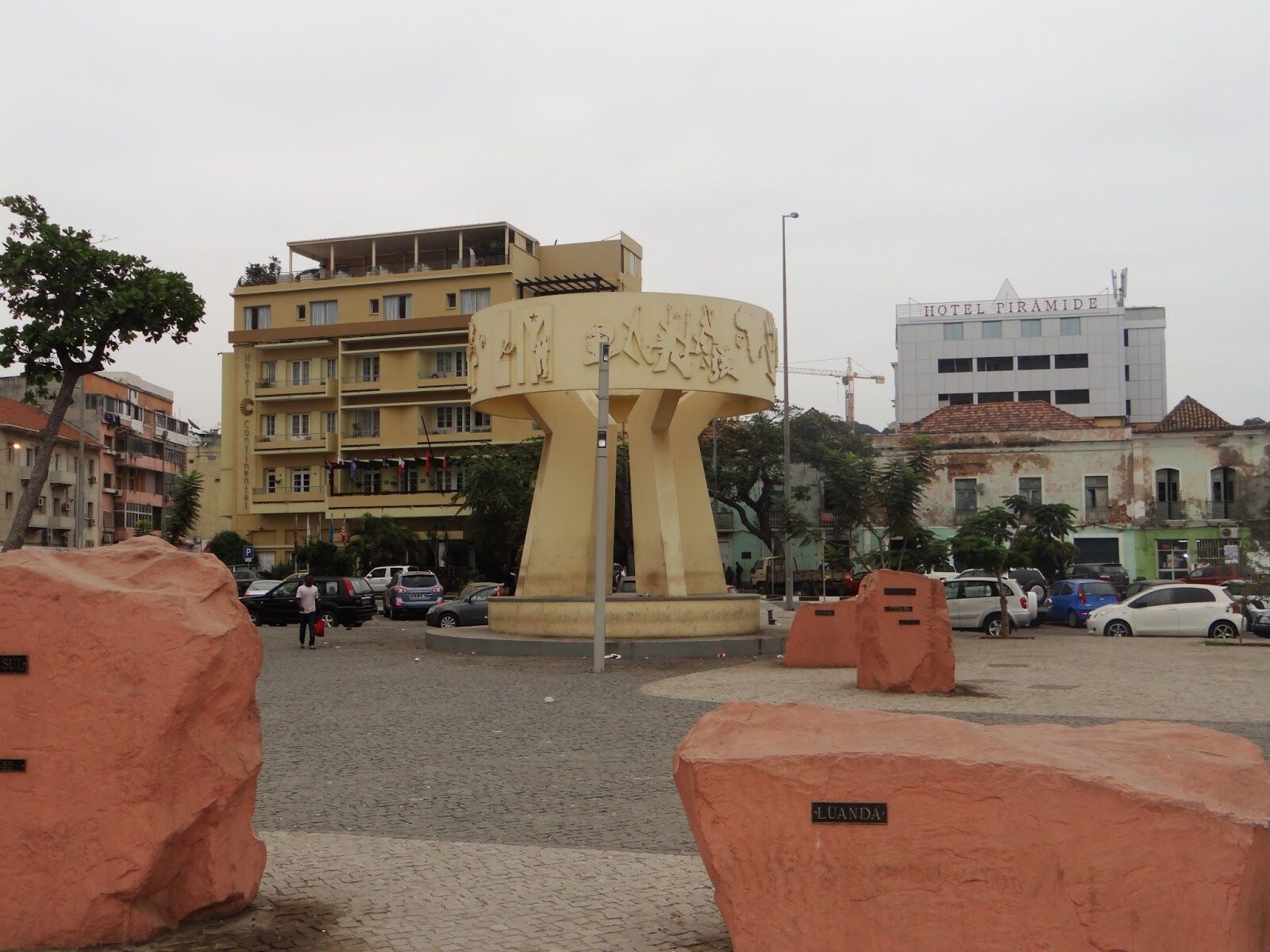 A large, beige, circular monument with inscriptions, surrounded by a paved area with orange barriers. In the background, there are multi-story buildings, including a hotel named "HOTEL PIRAMIDE".