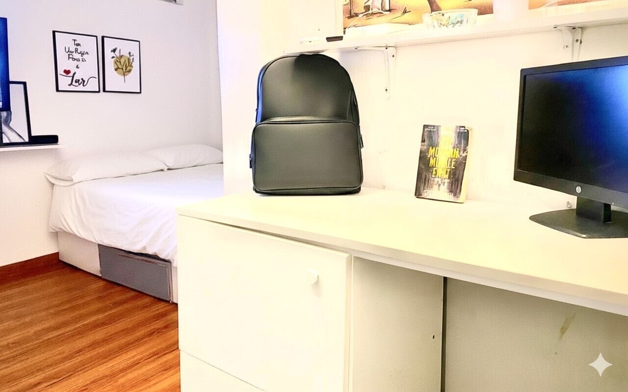 A bedroom with a bed, a desk with a computer monitor, a backpack, and framed pictures on the wall.