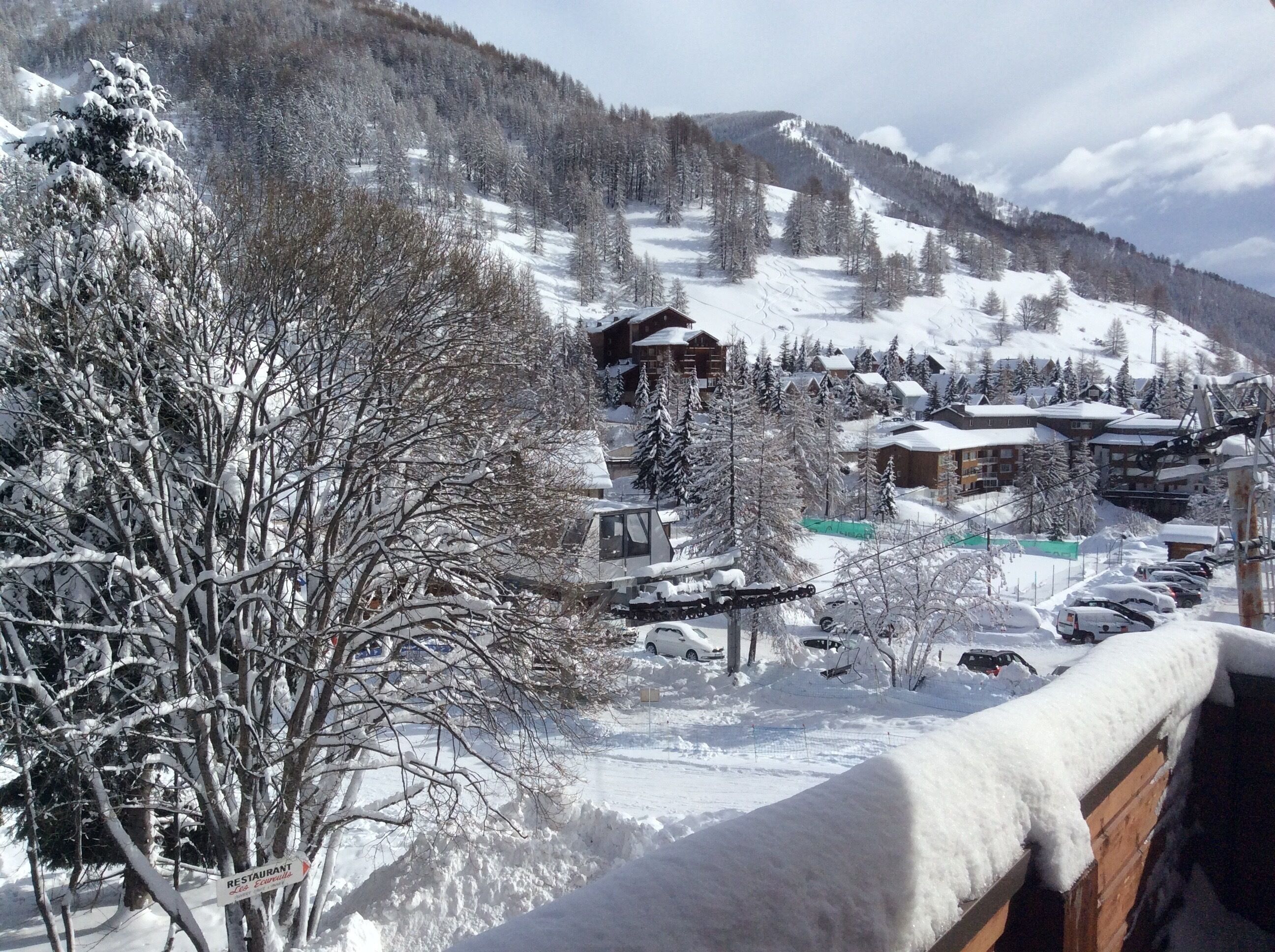 Un pueblo nevado en la montaña, con edificios, un telesilla y el anuncio de un restaurante.