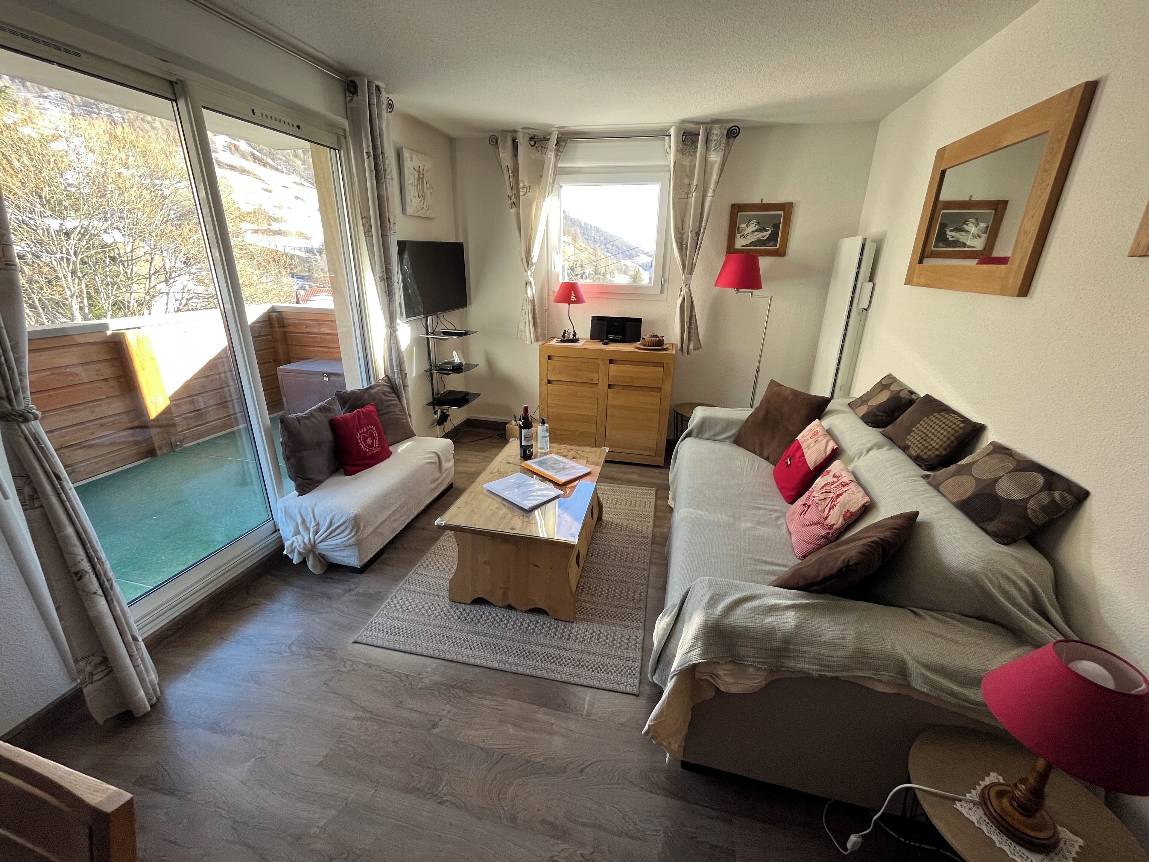 A cozy living room with a sofa, a coffee table, and a TV. There is a wooden cabinet and a red lamp. The room has large windows with curtains.