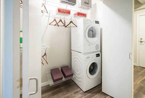 Un espace buanderie compact comprenant une machine à laver et un sèche-linge superposés, un étendoir et une petite étagère avec des boîtes.
