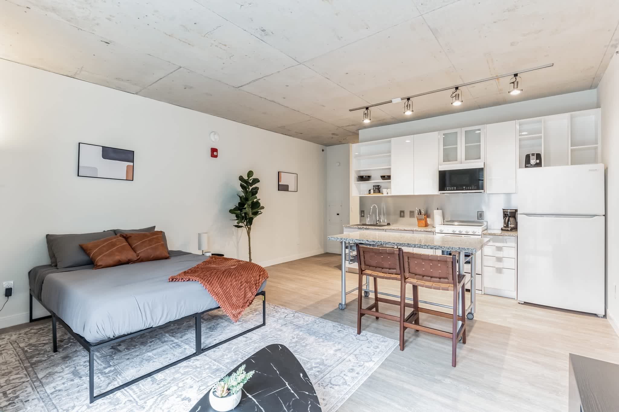 A modern apartment with a grey sofa, a small dining table, and a kitchen with white cabinets and appliances.