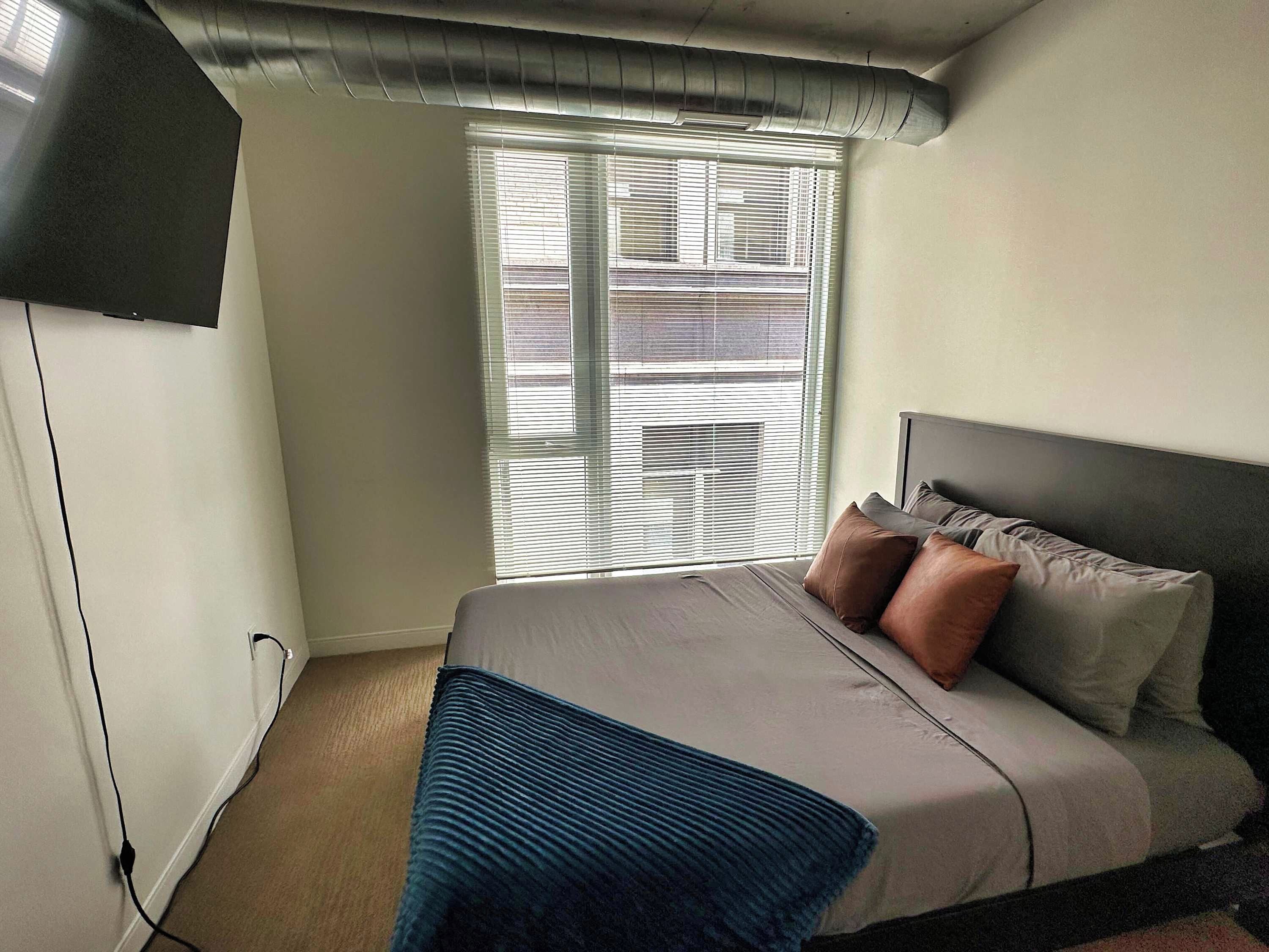 A bedroom with a bed, pillows, a TV mounted on the wall, and a window with blinds.