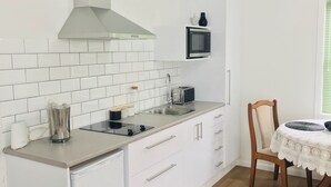 A modern kitchen with white cabinetry, a stainless steel range hood, a microwave, and a dining area with a wooden chair and a lace tablecloth.