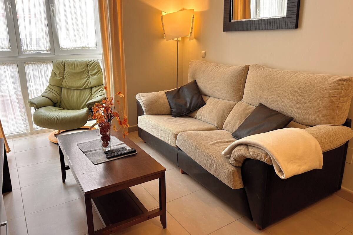 Una sala de estar con un sofá, una silla verde, una mesa con un jarrón y una lámpara.