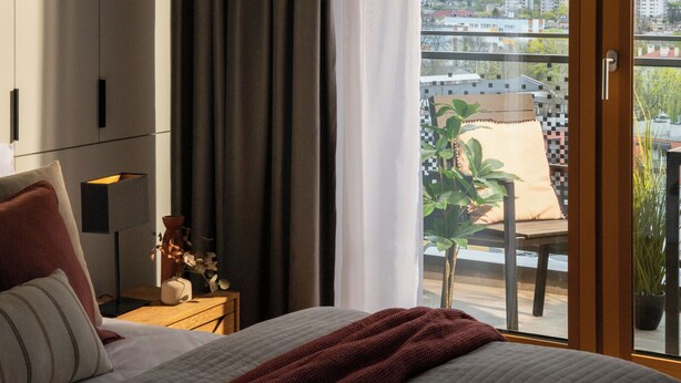 Un dormitorio con una cama, una mesita de noche, una lámpara, un ventanal con cortinas y vistas a la ciudad.
