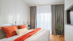 Una habitación de hotel moderna con una cama grande, cortinas grises, una planta en maceta y un televisor en la pared.