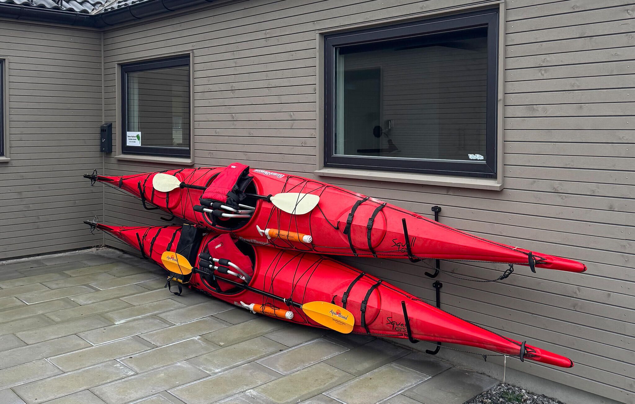 Two red kayaks with paddles and life jackets leaning against a building with windows.