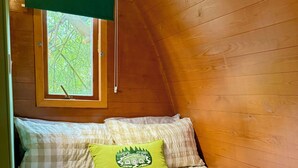 A cozy bedroom with a wooden ceiling, a bed with a green pillow, and a window with a view of trees. - Aelfred Pod (West Stow)