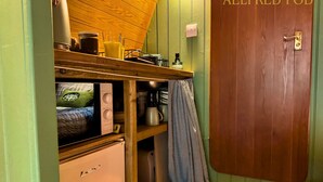 A compact kitchen with a refrigerator, a microwave, and a coffee maker. There's a wooden shelf above the counter holding a kettle and a glass. A checkered towel hangs on the door. - Aelfred Pod (West Stow)