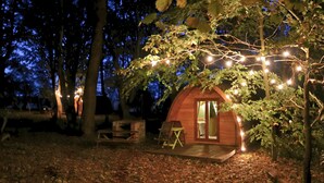 A small wooden cabin with a porch, surrounded by trees and illuminated by string lights. - Wulfrun Pod (West Stow)
