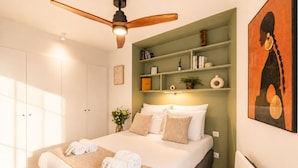 A bedroom with a bed, a ceiling fan, a bedside table with a lamp, a wall-mounted shelf with books and decorative items, and a painting on the wall. - 1530 Roux (Paris)