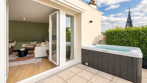 A modern outdoor living area with a hot tub, a sliding glass door leading to a furnished interior, and a view of a church spire in the background. - 1530 Roux (Paris)