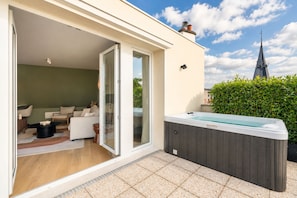 A modern outdoor living area with a hot tub, a sliding glass door leading to a furnished interior, and a view of a church spire in the background. - 1530 Roux (Paris)