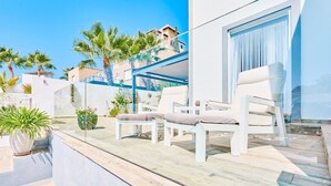 A modern outdoor seating area with white chairs and a glass balustrade overlooking a pool and a building with palm trees.