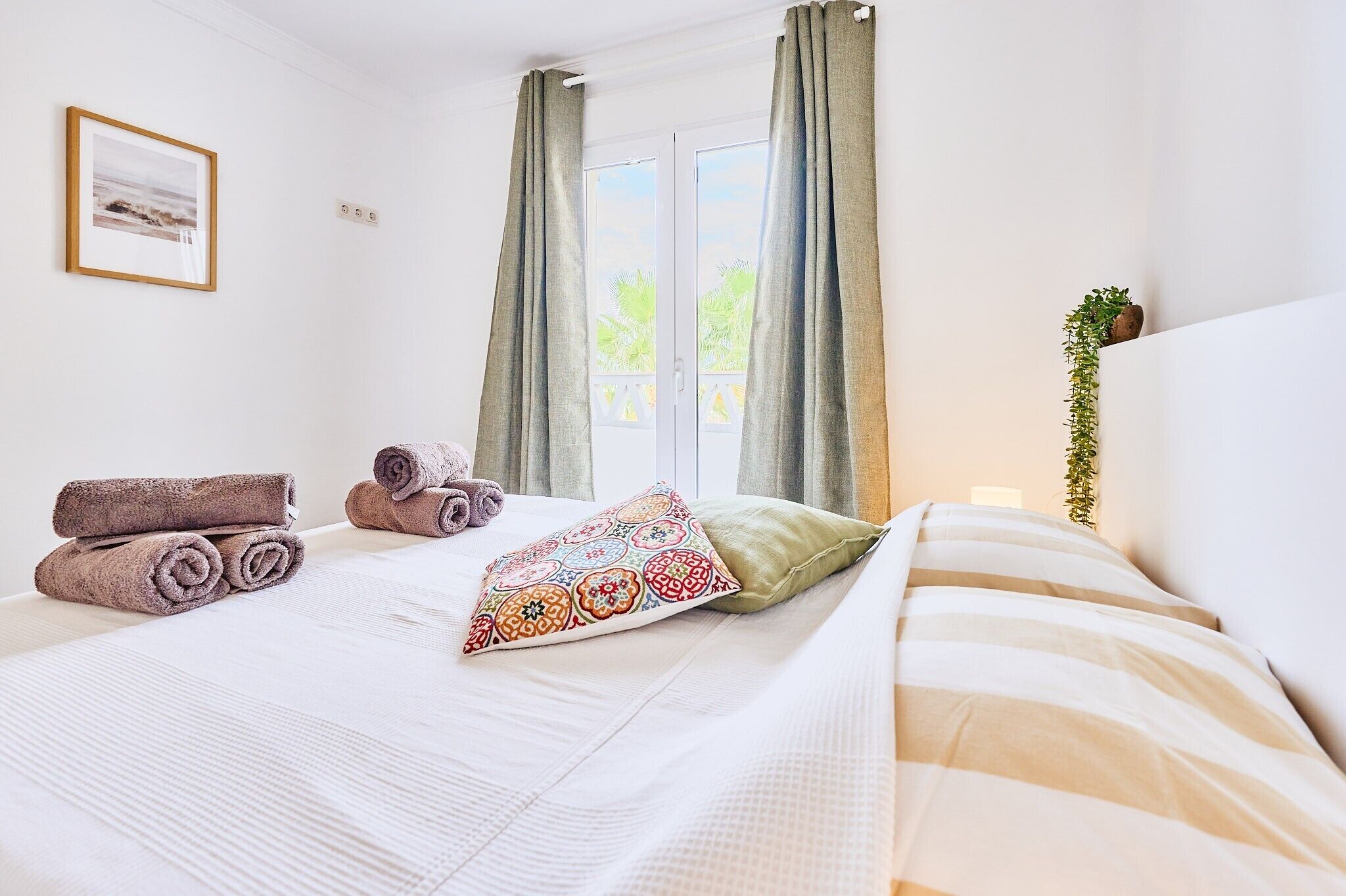 A hotel room with a bed, towels, a patterned pillow, a window with curtains, a wall-mounted picture, and a potted plant.